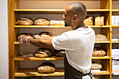 Back view of positive young African American male seller in apron smiling and taking bread from shelf while working in bakery shop