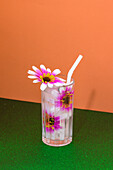 Transparent glass of refreshing cold drink decorated with pink flowers and straw placed on green surface against bright orange wall