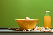 Glasflasche mit frischem Orangensaft und gelber Schale mit Popcorn auf glatter Oberfläche mit Popcornstreu vor grünem Hintergrund