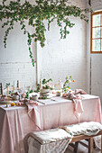 Eleganter Tisch mit Tellern und Blumen in der Nähe eines leckeren Kuchens auf einem rosa Tischtuch vor einer Backsteinwand