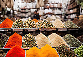 Reihen von gemischten farbigen pulverisierten Gewürzen und Samen in Schalen an einem lokalen Marktstand vor einem unscharfen, geschäftigen Markthintergrund