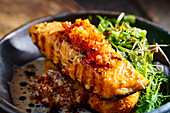 High angle of appetizing grilled salmon steak garnished with sauce and herbs served on black bowl placed on wooden cutting board