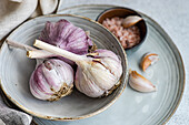 Raw garlic bulbs beautifully arranged on a ceramic plate with a bowl of salt accompanied by peeled cloves set against a soft textured backdrop