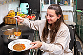 A smiling young woman in a wheelchair serves herself a healthy meal, showcasing independent living