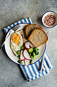 Top view of healthy lunch bowl with slices of bread, fried egg, fresh cucamelon, radish and tomato placed on a striped blue and white cloth with a small bowl of pink salt on the side, set against a gray textured backdrop