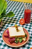 Appetizing healthy sandwich with fresh salad served on plate with olives near glass of tomato juice with glass straw on checkered tablecloth