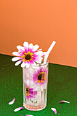 Transparentes Glas mit erfrischendem Kaltgetränk, verziert mit rosa Blumen und Strohhalm, auf grüner Fläche vor leuchtend oranger Wand