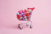 Composition of miniature shopping trolley cart with assorted multicolored sweets placed near fallen sweets on pink background