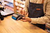 Crop anonymous male in casual clothes standing at wooden table while operating and processing financial payment on POS machine in shop