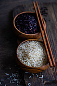 High angle of Raw wild black rice and peeled white rice in the bowls with chopsticks placed on cutting board on wooden table