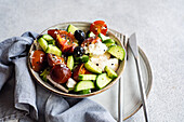 High angle of ceramic bowl with tasty healthy salad and vegetables placed with sliced cucumber and tomato with cutlery and fabric over gray table