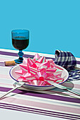 Generative AI image of a whimsical Valentine's day setup on a striped tablecloth featuring a glass, a plate with a giant pink bow, and a napkin.