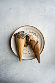 Top view of waffle cones with coffee and chocolate ice cream with multicolored sugar sprinkles and nuts on top in a ceramic dish