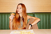 Young woman with red hair and piercing sitting at table and enjoy eating hamburger in restaurant