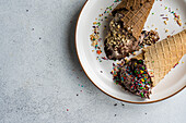 Top view of waffle cones with coffee and chocolate ice cream with multicolored sugar and nuts on a ceramic plate