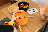 High angle of anonymous women while sitting at table and carving with tool scary teeth on orange pumpkin and preparing for Halloween decoration in day