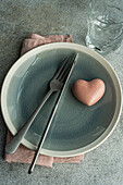 Top view of ceramic plate with cutlery and napkin with heart shaped decor crystal placed on concrete surface at kitchen table for meal during Valentine's day celebration