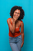Excited African American female model with Afro hairstyle in casual outfit laughing at joke with closed eyes and covering face against blue background