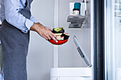 Seitenansicht eines anonymen männlichen Kochs in Schürze und umgeschlagenen Ärmeln, der im Stehen geschnittene Wassermelonenfrüchte aus einer roten Schüssel in einen automatischen Mülleimer wirft
