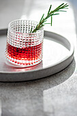 Elegant cocktail with cherry and apple juice mixed with vodka garnished with a fresh rosemary sprig presented on a circular tray