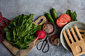 Draufsicht auf eine Reihe frischer Zutaten wie Spinat, Tomaten, Zucchini und Gewürze auf einer strukturierten Oberfläche, ergänzt durch Holzutensilien und eine rustikale Schere