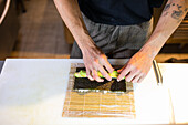High angle of anonymous tattooed male in apron standing at wooden table and arranging avocado slices on seaweed wrap placed on bamboo mat in blurred kitchen