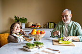 Smiling elderly grandfather with eyeglasses cutting lettuce and looking at cute granddaughter while sitting together at table at home