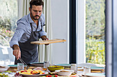 Bearded male private chef in apron putting appetizer on plate with tongs while serving delicious dishes for breakfast in light dining room