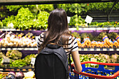 Back view of unrecognizable female customer with backpack and shopping basket choosing fresh vegetables on shelves at at supermarket