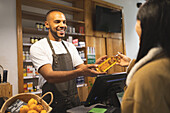 Positive young African American male seller in apron smiling and giving bottle of oil to customer while standing at counter during work
