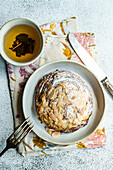 Top view of sweet almond pastry on napkin with fork and knife with cup of green tea against gray background in sunlight