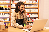 Positive young male in casual clothes with apron smiling and looking at screen while talking on smartphone and browsing laptop over table in drugstore