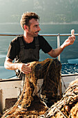 Ganzkörperaufnahme eines lächelnden männlichen Fischers, der beim traditionellen Fischfang Muscheln aus dem Netz fischt, vor einer Meereslandschaft in Soller auf der Baleareninsel Mallorca