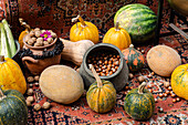 A colorful autumnal arrangement featuring a variety of pumpkins, a melon, hazelnuts in a rustic pot, walnuts adorned with a pink flower, and fresh corn cobs, all set on an ornate, patterned carpet.