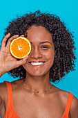 Happy young Hispanic female with curly hair covering eye with slice of orange and looking at camera against blue background