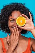 Happy young Hispanic female with curly hair covering eye with slice of orange and covering mouth with hand while looking at camera against blue background