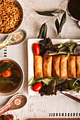 A top view of a traditional Asian meal featuring crispy spring rolls alongside soup and noodles, beautifully arranged on a patterned tablecloth.