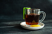 A warm cup of tea with a green tea infuser and lemon slices on a saucer, placed on a dark textured table