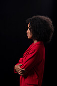 Side view of serious young Hispanic female model with Afro hairstyle in red coat standing arms crossed against black background