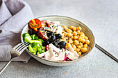 Hoher Winkel einer Schüssel mit gesundem Salat und Gemüse mit Kichererbsen, Sesam und Oliven mit Besteck und Stoff auf einem hellgrauen Tisch