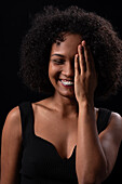 Shy young Hispanic female with curly hair covering half face and smiling happily against black background