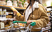 Crop unrecognizable female customer in casual outfit standing near shelves with shopping trolley and choosing products during sale in supermarket
