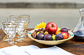 A wooden table displaying an array of fresh fruits in a bowl, accompanied by empty wine glasses and a pitcher of juice, set against a muted backdrop.