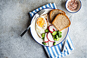 Draufsicht auf eine gesunde Mittagsschüssel mit Brotscheiben, Spiegelei, frischer Gurke, Rettich und Tomate auf einem blau-weiß gestreiften Tuch, daneben eine kleine Schale mit rosa Salz, vor einem grau strukturierten Hintergrund