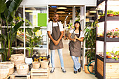 Young African American positive saleswomen in aprons smiling and looking at camera as they stand by a glass door surrounded by plants