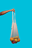 Crop anonymous hand of person holding eco friendly shopping bag with fresh fruits against blue background