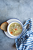 From above of warm bowl of chicken and vegetable soup with a side of whole grain bread displayed on a textured grey background with a striped napkin and a spoon