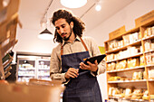 From below portrait of serious ethnic male worker with long curly hair in casual clothes standing near shelves and working on tablet in modern lighted grocery shop