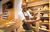 Happy young African American male seller in apron putting fresh baked bread into paper bag while standing near shelves during work process
