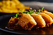 Delicious fried breaded meat garnished with mushrooms and herbs served on black plate near French Fries against blurred background